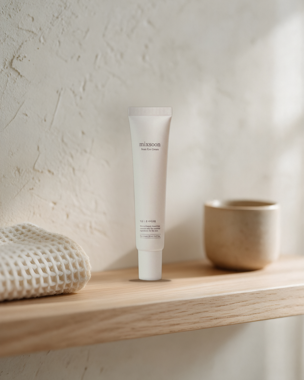 Mixsoon Bean Eye Cream on a wooden bathroom shelf beside a waffle towel and a ceramic cup
