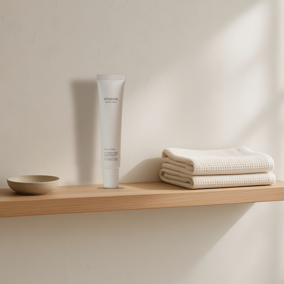 Mixsoon Bean Eye Cream on an oak shelf between a stone dish and folded waffle towels